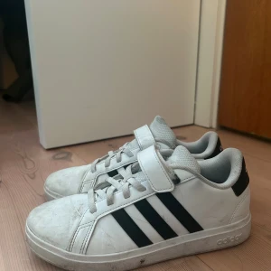 Vita Adidas sneakers med svarta ränder - Klassiska vita Adidas sneakers med svarta ränder på sidorna och kardborreband upptill. Skorna har rund tå, snörning och platt sula med bikakemönster undertill. Ovandelen är i syntetmaterial och textil, perfekt för en sportig och avslappnad stil.
