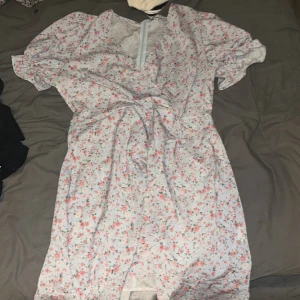 Blommig kortklänning från Chiquelle - Supersöt kortklänning från Chiquelle i ljusblått med små blommor i rosa och grönt. Klänningen har v-ringning, korta puffärmar och knyt i midjan. Tillverkad i 100% polyester och har dragkedja i ryggen. Perfekt för sommarens alla tillfällen!