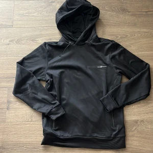 Svart hoodie från Sail Racing - Svart hoodie från Sail Racing med diskret logga på bröstet. Tröjan har huva med snörning och långa ärmar. Tillverkad i ett slätt, syntetiskt material som känns sportigt och modernt. Perfekt för dig som gillar stilren och enkel design.