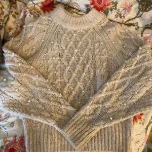Beige stickad tröja med pärlor - Superfin beige stickad tröja med flätstickat mönster och pärldetaljer på ärmar och framstycke. Tröjan har hög hals och långa ärmar med breda muddar. Perfekt för dig som gillar mysiga och snygga plagg med det lilla extra.