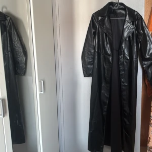 Svart lång skinnrock - Säljer en svart lång skinnrock med klassisk krage och knäppning framtill. Jackan har en rak passform och långa ärmar, perfekt för dig som vill ha en edgy och stilren look. Materialet är glansigt och ger en cool Matrix-vibe.