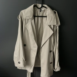 Beige trenchcoat från hm - Stilren beige trenchcoat från Iconic med klassisk krage, knappar och bälte i midjan. Jackan har långa ärmar med knäppning vid ärmslut och axelklaffar. Perfekt för dig som vill ha en tidlös och snygg look.