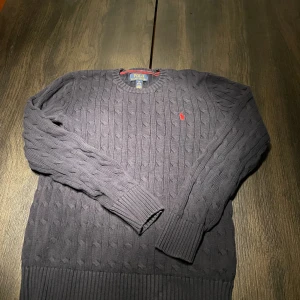 Mörkblå stickad tröja Polo Ralph Lauren - Kabelstickad mörkblå tröja från Polo Ralph Lauren med rund halsringning och röd broderad logga på bröstet. Tröjan har långa ärmar och breda ribbade muddar vid ärmslut och nederkant. Perfekt för dig som gillar klassisk och stilren look.