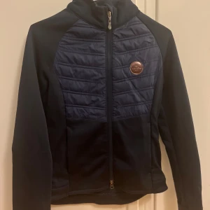 Marinblå ridjacka från JH Collection - Snygg marinblå ridjacka/vindjacka från JH Collection med quiltad front och dragkedja hela vägen. Jackan har hög krage, lång ärm och logga på bröstet samt tryck på ryggen. 