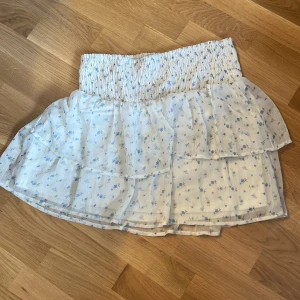 Blommig volangkjol från Vero Moda - Supersöt kort kjol från Vero Moda i vitt med små blå blommor. Kjolen har bred smock i midjan och flera lager volanger som ger en luftig och romantisk känsla. Perfekt för varma dagar och lätt att matcha med en enkel topp.