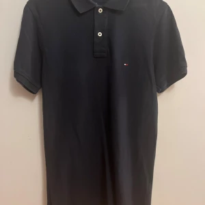 Svart pikétröja Tommy Hilfiger S - Svart pikétröja från Tommy Hilfiger i slim fit-modell. Klassisk krage med två knappar och diskret logga broderad på bröstet. Tillverkad i bomull och har korta ärmar. 