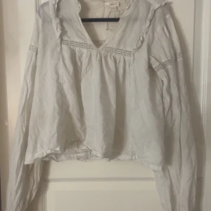 Vit blus med volang från Gina Tricot - Säljer en luftig vit blus från Gina Tricot med v-ringning, volangdetaljer vid halsen och dekorativa spetsinslag på ärmar och över bröstet. Blusen har långa ärmar och en avslappnad passform, perfekt för en chill och stilren look.