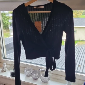 Svart omlottopp från NA-KD, S - Svart croppad omlottopp från NA-KD med långa ärmar och knyt i midjan. Tunt ribbat material som ger en lätt transparent känsla och markerad v-ringning. Perfekt för dig som gillar trendiga och stilrena plagg med snygg passform.