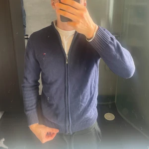 Marinblå full zip kofta från Tommy Hilfiger - Säljer en marinblå kofta från Tommy Hilfiger med dragkedja framtill och ribbade muddar. Klassisk liten logga på bröstet och hög krage med kontrastfärg på insidan. Perfekt till lager-på-lager.