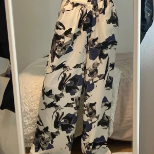 Vida mönstrade byxor i vitt och svart - Säljer ett par riktigt snygga vida byxor med abstrakt svart och mörkblått mönster på vit botten. Byxorna har hög midja och är tillverkade i ett mjukt, flowy material som känns luftigt och skönt. Perfekta för dig som gillar statement-plagg och vill sticka ut.