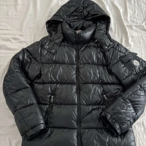 Svart dunjacka från Moncler - Snygg svart pufferjacka från Moncler med glansig finish och huva. Jackan har en dragkedja framtill och ett broderat Moncler-märke på ärmen. Perfekt för kalla dagar.