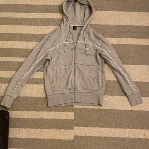 Grå HUGO BOSS hoodie med dragkedja  - Grå hoodie från H&M med hel dragkedja, huva med snörning och två stora fickor framtill. Tröjan har vita detaljer längs ärmarna och ribbade muddar vid ärmslut och nederkant. Perfekt för en avslappnad stil.