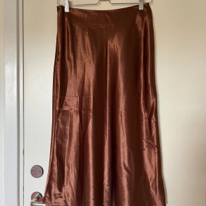 Brun satinkjol - Snygg midikjol i skimrande brunt glansigt tyg, lätt utställd passform. Materialet ger ett lyxigt intryck och faller snyggt längs benen. Midjemått 72 cm, passar s/m. Dragkedja i sidan. Köpt second hand. Fint skick! Säljer en liknande i orange också! 