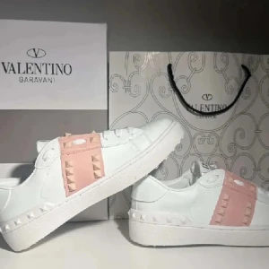 Valentino Garavani Rockstud sneakers - Snygga vita Valentino Garavani Rockstud sneakers med rosa detaljer och ikoniska nitar. Skorna har rund tå, platt vit sula och är tillverkade i skinn. Perfekta för dig som vill sticka ut med en lyxig och trendig look.