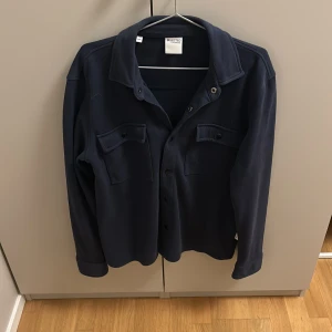 Mörkblå overshirt från Selected Homme - Snygg mörkblå overshirt från Selected Homme med klassisk krage och två bröstfickor med lock och knappar. Rak passform och långa ärmar, perfekt att bära öppen eller stängd. Stilren och enkel design som funkar till många outfits.