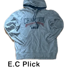 Champion hoodie - Grå hoodie från Champion med blått och rött tryck på bröstet där det står 'CHAMPION New York 1919'. Tröjan har huva med snörning, känguruficka och långa ärmar. Perfekt för en avslappnad och sportig stil.