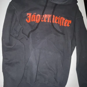 Svart Jägermeister hoodie med tryck - Svart hoodie från Jägermeister med orange logga och text både fram och bak. Stort tryck med hjort och kors på ryggen. Klassisk känguruficka och huva med snörning. Perfekt statement-plagg för dig som gillar streetwear och snygga loggor. Använd ca 3-4 gånger utan defekter.