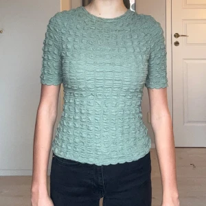 Grön ribbad topp med struktur - Säljer en ljusgrön topp med korta ärmar och rund hals. Toppen har ett ribbat, rutigt strukturmönster som ger en cool och unik look. Passformen är slim och materialet känns mjukt och stretchigt, perfekt till jeans eller kjol.