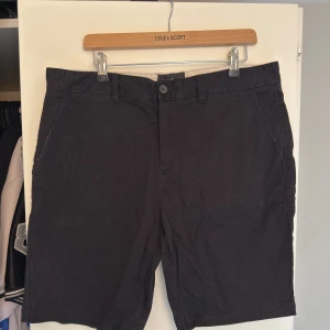 Svarta shorts från Lyle & Scott W36 - Klassiska svarta shorts från Lyle & Scott i bomull med normal passform. De har bälteshällor, knapp och dragkedja framtill samt två fickor bak, varav en med den ikoniska gula loggan. Perfekta för en stilren och enkel look.
