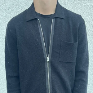 Svart kofta med dragkedja - Svart kofta med lång ärm och silverfärgad dragkedja framtill. Klassisk krage och en bröstficka på vänster sida. Mjuk och lite tjockare känsla, perfekt för lager-på-lager. Enkel och stilren design som funkar till det mesta.