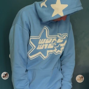 FET Blå hoodie med stjärnor och tryck (streetwear) - Säljer en ljusblå hoodie från World Wide med stora vita stjärnor på huvan och ett coolt vitt tryck framtill. Tröjan har huva, känguruficka och långa ärmar. Perfekt för dig som gillar streetwear och vill sticka ut med en unik design.