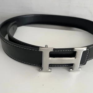 Svart H skärp med H-spänne - Snyggt svart skärp från  med ikoniskt H-spänne i silverfärgad metall. Bältet är gjort i äkta skinn med vita kontrastsömmar längs kanterna. Perfekt accessoar för att lyfta din outfit med en lyxig touch.