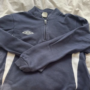 Mörkblå Umbro half zip sweatshirt XS - Sportig mörkblå sweatshirt från Umbro i storlek XS. Tröjan har vitt Umbro-logo broderat på bröstet, vita paneler på sidorna och en praktisk half zip-dragkedja. Långärmad modell i mjuk bomullsblandning, perfekt för träning eller chill.
