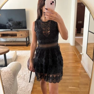 Svart GUESS spetsklänning med blommönster - Supersnygg svart kortklänning med blommig spets och transparenta detaljer i midjan från Guess. Klänningen är ärmlös och har en rundad halsringning med fina spetskanter. Kjolen är lätt utställd och ger en feminin siluett. Perfekt för dig som vill sticka ut med en elegant look.