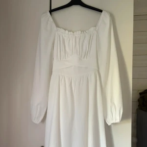 Vit kortklänning med rosett och långa ärmar - Superfin vit kortklänning med långa puffärmar och fyrkantig ringning. Klänningen har en söt rosett i ryggen och är gjord i ett lätt, strukturerat tyg som känns luftigt. Vet tyvärr ej varumärke då den är köpt här på Plick men passade inte mig och lappen är bortklippt🎀