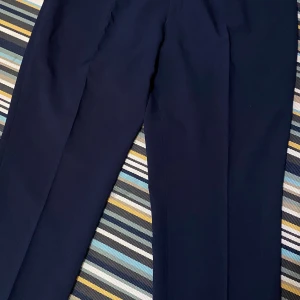 Mörkblå kostymbyxor med dragkedja - Snygga mörkblå kostymbyxor med klassisk rak passform. Byxorna har dragkedja och hake i midjan samt diskret knapp. Perfekta för en clean och stilren look. Materialet känns mjukt och följsamt, troligtvis polyester eller bomullsblandning. Midjan är 112 cm och längden är 102 cm