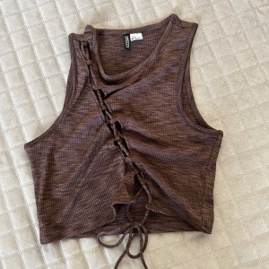 Brun ribbad croppad topp H&M Divided - Säljer en brun croppad linnetopp från H&M Divided i ribbat material. Toppen har snörning framtill som ger en edgy look och är ärmlös med rund halsringning. Perfekt för dig som gillar trendiga detaljer och vill sticka ut lite extra.