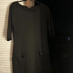 Svart långklänning från Filippa K - En stilren svart långklänning från Filippa K med trekvartsärm och två stora fickor framtill. Klänningen har en rund halsringning och är tillverkad i mjuk bomull, perfekt för en clean och minimalistisk look.