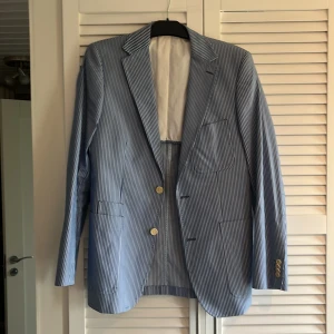 Blåvit randig kavaj från Morris - Snygg blåvit randig kavaj från Morris med klassisk passform. Kavajen har två ljusa knappar framtill och tre dekorativa knappar vid ärmslutet. Fodret är vitt med röda detaljer och märkesetikett. Perfekt för dig som gillar stilrena och fräscha looks. Kaffe fläck på inte försökt få bort de så säljs billigt