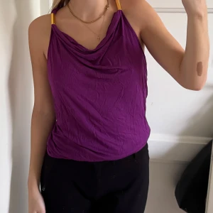 Lila linnetopp med gulddetaljer - Säljer en snygg lila linnetopp med draperad halsringning och smala axelband med guldfärgade detaljer. Toppen har öppen rygg med en guldfärgad ring som samlar banden. Perfekt för dig som gillar färg och unika detaljer.