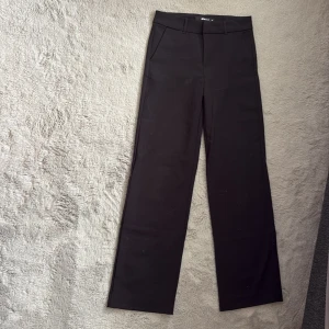Svarta kostymbyxor Gina Tricot 32 - Stilrena svarta kostymbyxor från Gina Tricot i storlek 32. Byxorna har hög midja, raka ben och hällor för bälte. Klassisk design med fickor fram och en fuskficka bak. Perfekta för en clean och snygg look.