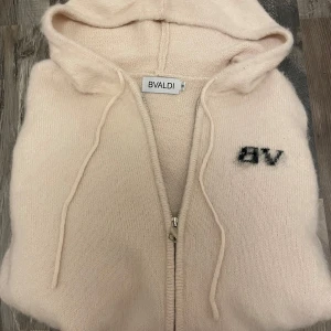 Beige stickad hoodie från BVALDI - Mysig beige hoodie från BVALDI i stickat material med huva och snörning. Tröjan har dragkedja framtill, två fickor och svart BV-brodyr på bröstet. Perfekt för chill dagar och snygg till jeans eller mjukisbyxor.