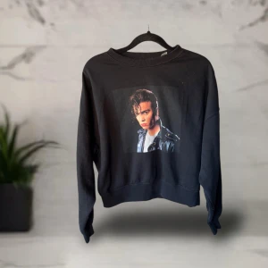 Svart Cry-Baby sweatshirt med tryck - Svart sweatshirt med stort tryck av Cry-Baby på ryggen och en cool bild på framsidan med läderjacka och vit t-shirt. Tröjan har en loose passform, ribbade muddar och är långärmad. Perfekt för dig som gillar film och retrostil.