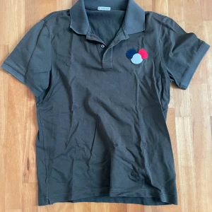 Svart Moncler pikétröja med brodyr - Svart pikétröja från Moncler med klassisk krage och knappslå. På bröstet finns en broderad detalj i rött, blått och vitt. Tröjan är tillverkad i bomull och har korta ärmar. Perfekt för en clean och stilren look.