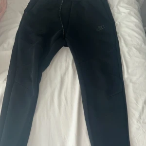 Svarta mjukisbyxor från Nike - Svarta mjukisbyxor från Nike med diskret logga på låret. Byxorna har dragsko i midjan och är tillverkade i mjukt material som känns skönt mot huden. Perfekt för chill eller träning. Snygg, enkel design med smal passform och praktiska fickor.