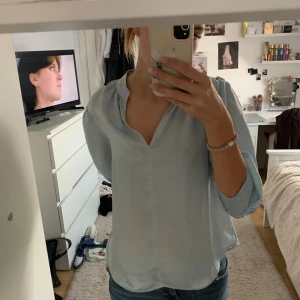 Ljusblå blus med v-ringning - Säljer en ljusblå blus med v-ringning och trekvartsärmar. Blusen har lätt puffade axlar och smockdetalj vid ärmsluten. Den är luftig och har en avslappnad passform, perfekt till jeans eller kjol.