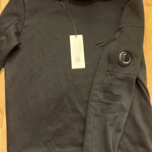 C.P. Company tröja - Säljer en stilren svart långärmad tröja från C.P. Company. Tröjan har en unik detalj på ärmen och är perfekt för en modern look. Märkets logga finns både på etiketten och som en snygg detalj. Köpt på CareOfCarl, kvitto finns. Nypris 2199kr