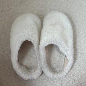Vita fluffiga tofflor - Mysiga vita tofflor med fluffig yta och mjuk insida. De har en enkel slip-in design och platt sula, perfekta att glida i hemma. Utsidan är täckt av syntetiskt pälsmaterial som ger en riktigt cozy känsla.