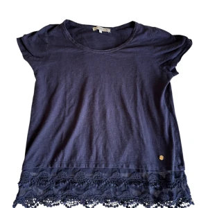 Mörkblå topp med spetskant - Supersöt mörkblå topp från Odd Molly med korta ärmar och rund halsringning. Nedtill finns en bred, genombruten spetskant med broderade detaljer och små bollar. Toppen är i mjuk bomull och har en liten guldfärgad logga vid fållen.
