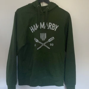 Grön Hammarby hoodie med tryck - Mörkgrön hoodie från Hammarby med vitt tryck framtill, snörning i huvan och stor magficka. Klassisk sportig stil med långa ärmar och mjukt material, perfekt för dig som gillar supporterkläder. 