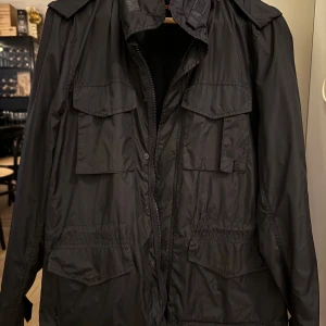 Svart vindjacka från Aspesi - Svart vindjacka från Aspesi i nylon med flera stora fickor framtill och dragkedja. Jackan har hög krage, justerbar midja och en minimalistisk design. Perfekt för dig som gillar stilrena och funktionella plagg. Broderad Aspesi-logga på insidan.