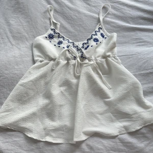 Vit broderad linnetopp med knyt - Supersöt vit linnetopp med tunna axelband och blå broderade blommor vid bysten. Toppen har v-ringning, knyt i midjan och är tillverkad i ett lätt, strukturerat tyg. Perfekt för varma dagar och ger en fräsch och somrig vibe. Tröjan är från shein. 