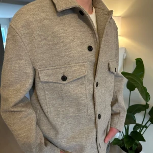 Beige overshirt från Zara i ullmix - Stilren beige/grå overshirt från Zara i mjuk ullmix. Jackan har klassisk krage, svarta knappar framtill och två stora bröstfickor. Normal passform. 
