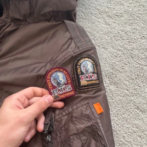Brun Parajumpers vinterjacka med päls - Säljer en brun Parajumpers vinterjacka med huva och äkta pälsdetalj. Jackan har flera fickor, ikonisk gul rem vid kragen och broderade PJS-patchar. Fodrad insida för extra värme och dragkedja med knappar framtill. Perfekt för kalla dagar.