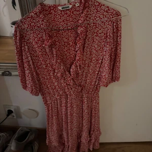 Röd blommig klänning från Indiska - Supersöt röd klänning från Indiska med vitt blommönster, v-ringning och volangdetaljer. Klänningen har korta ärmar och en flowy passform som ger en avslappnad men ändå feminin look. Perfekt för varma dagar och sommarhäng!