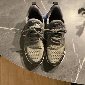 Nike Air 270 grå sneakers  - Nike Air 270 sneakers i grått med svarta och blå detaljer. Ovandel i mesh för bra andningsförmåga, snörning framtill och synlig blå air-sula baktill. Snygg sportig look och bekväm passform, perfekt för dig som gillar streetstyle.